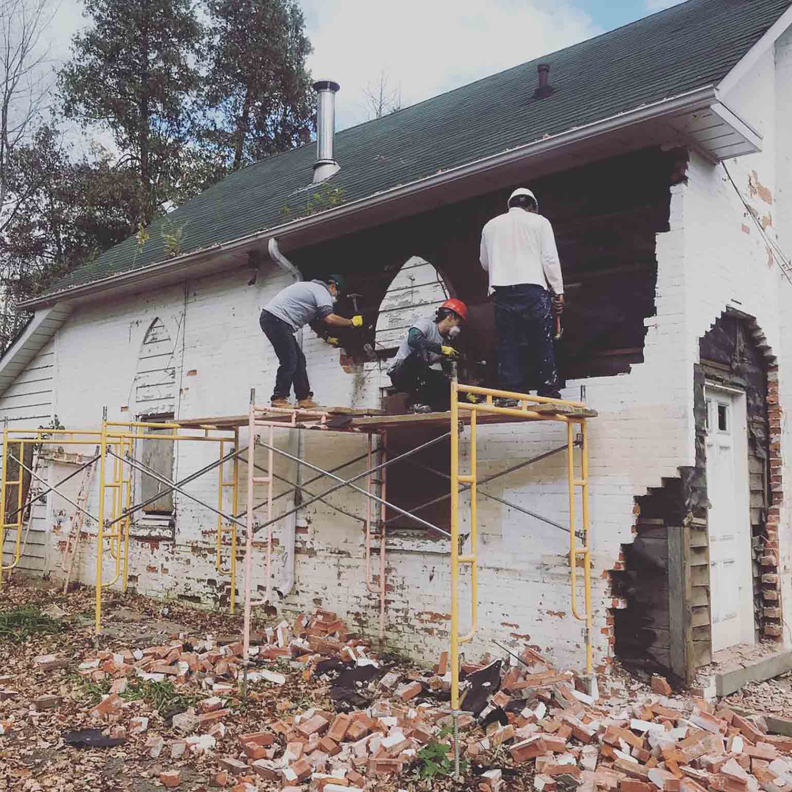 Three workers help demolish a white brick wall for a home renovation to help the homeless.