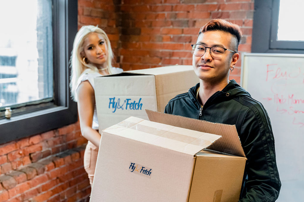 A man and a woman holding shipping boxes printed with the Fly and Fetch logo. 