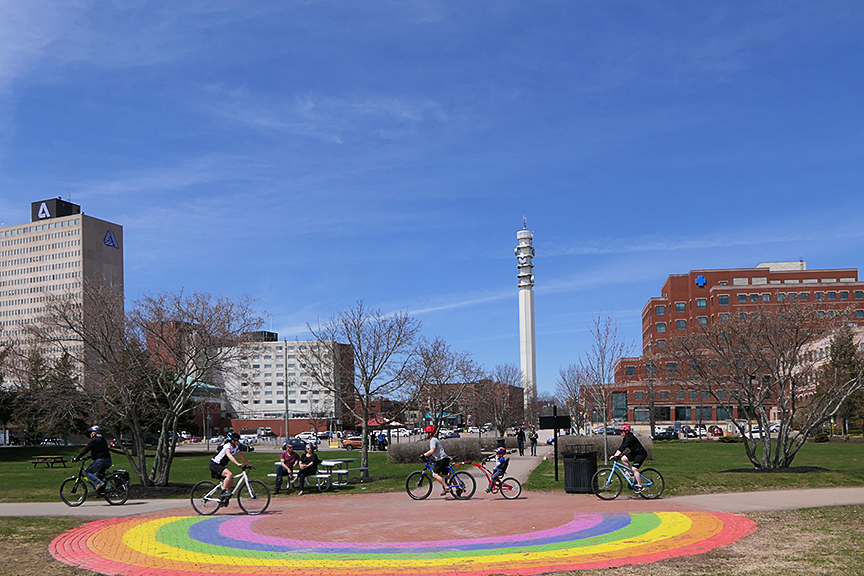 A park plaza on a sunny day in downtown Moncton with young people bicycling, and a rainbow painted on the sidewalk.