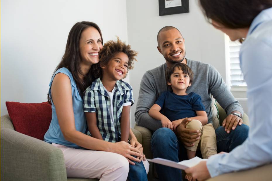 A young family of four with two sons sitting on a white couch, smiling while a mortgage specialist discusses moving mortgages for their home