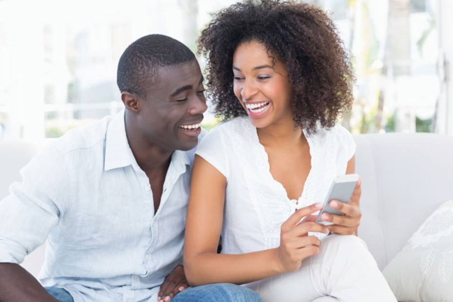 A picture showing a happy couple sitting on a sofa, laughing at something on her phone.