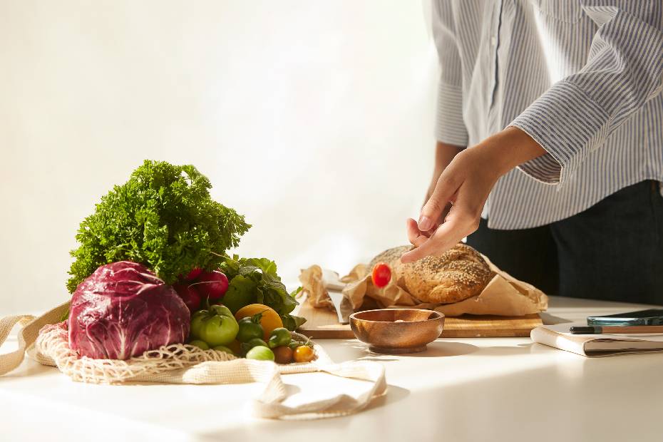 A hand places a cherry tomato into a bowl on a table of vegetables and bread
