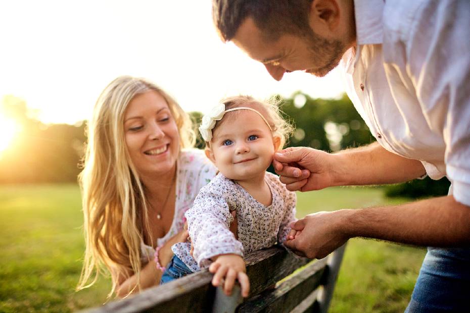 A picture showing a young happy family sitting on a bench in the park, with mom holding the baby girl and dad touching the baby's face.