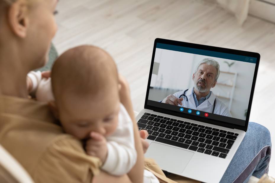 Image shows a mother holding baby, using laptop, making video call to pediatrician