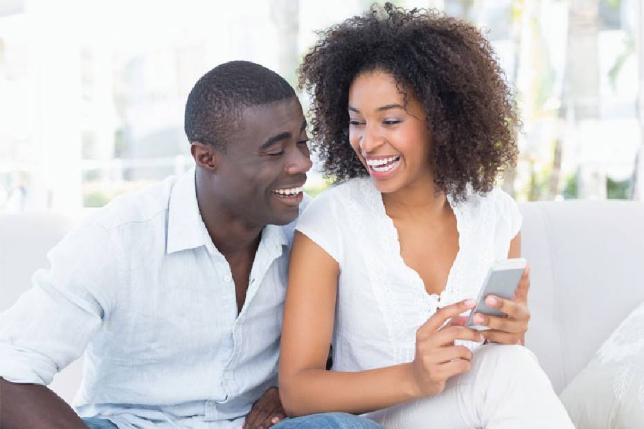 A picture showing a happy couple sitting on a sofa, laughing at something on her phone.
