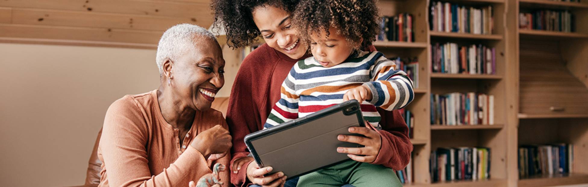 A grandmother smiles and sits with her daughter and granddaughter while looking at a tablet together.