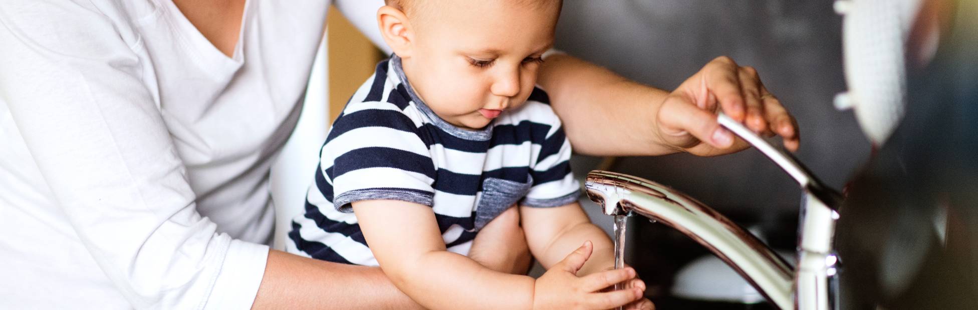 A parent holds their baby at the kitchen sink with the faucet running