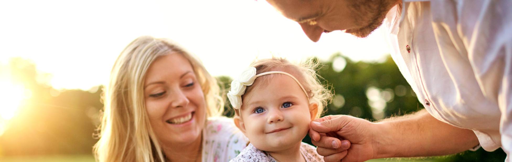 A picture showing a young happy family sitting on a bench in the park, with mom holding the baby girl and dad touching the baby's face.