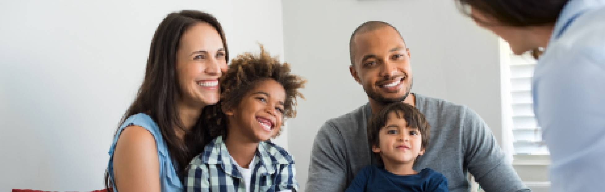 A young family of four with two sons sitting on a white couch, smiling while a mortgage specialist discusses moving mortgages for their home