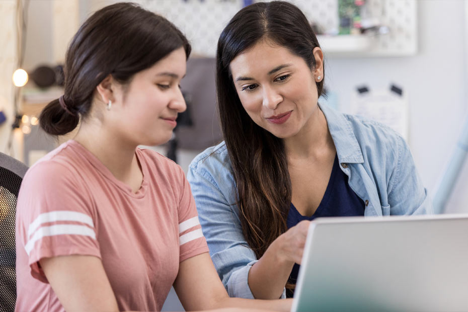 A young mother and teenage daughter in a bright kitchen using a laptop to learn about spending, saving, and credit in order to help her understand her finances.
