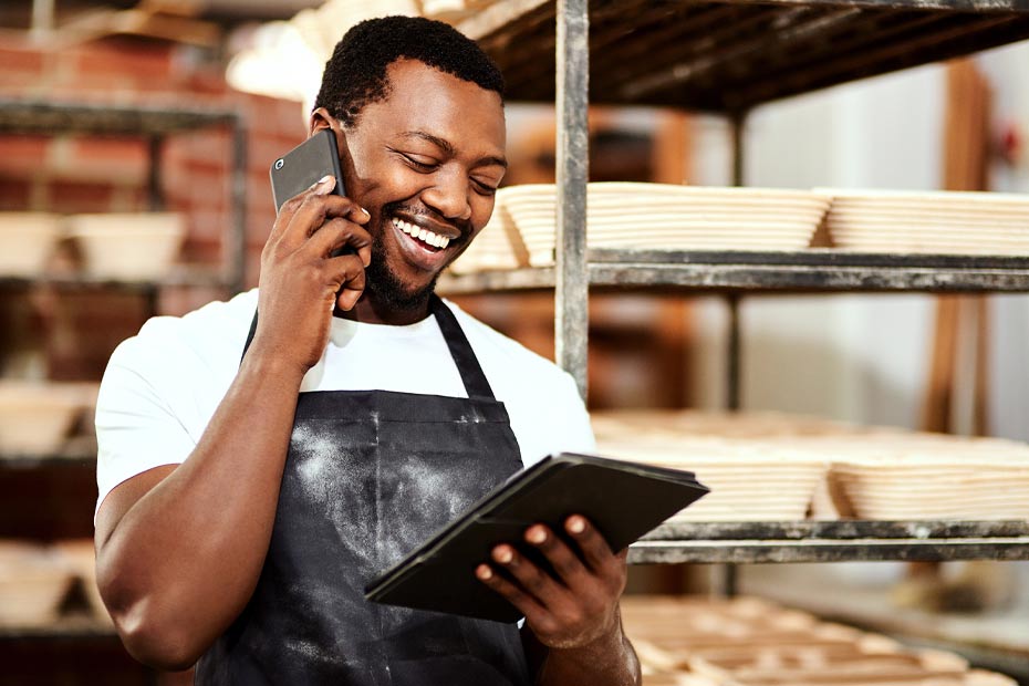 A smiling business owner in his stockroom, making a payment on his tablet while on the phone