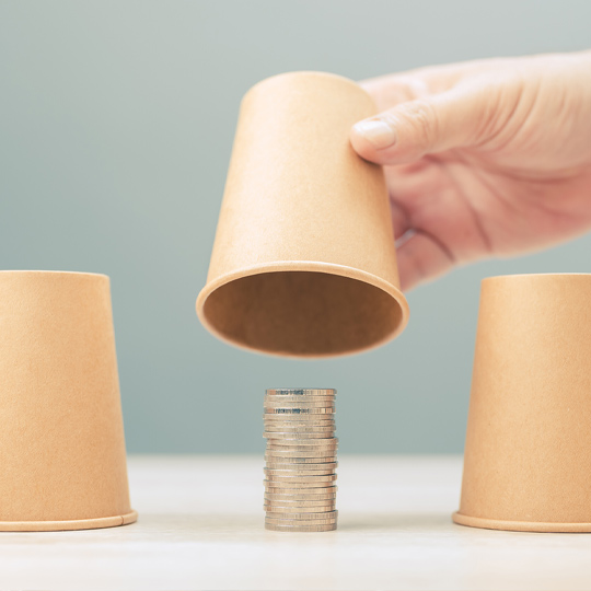 Image of 3 cups facing down and a hand raising the middle cup, revealing a stack of coins.