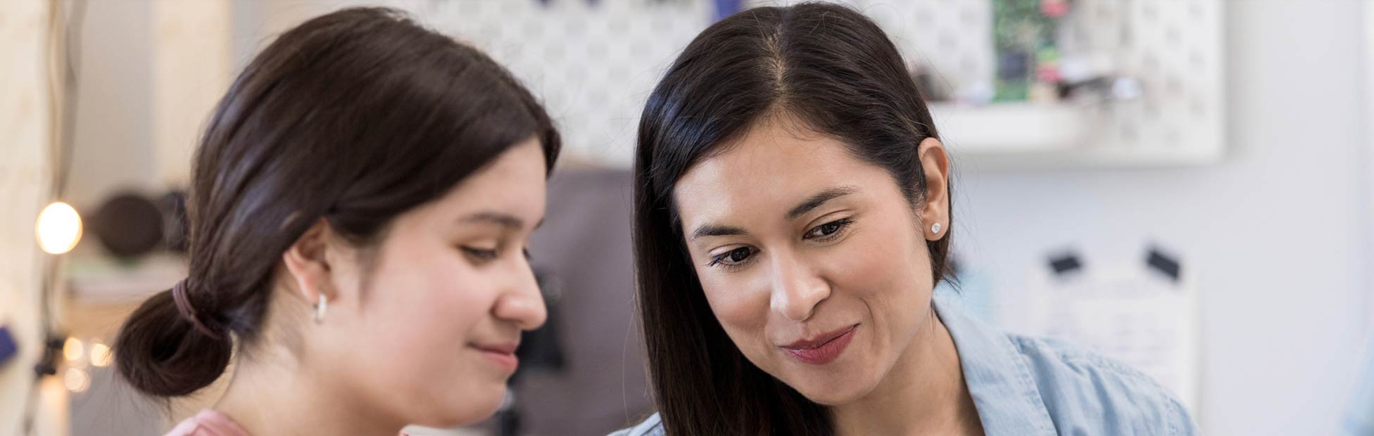 A young mother and teenage daughter in a bright kitchen using a laptop to learn about spending, saving, and credit in order to help her understand her finances.