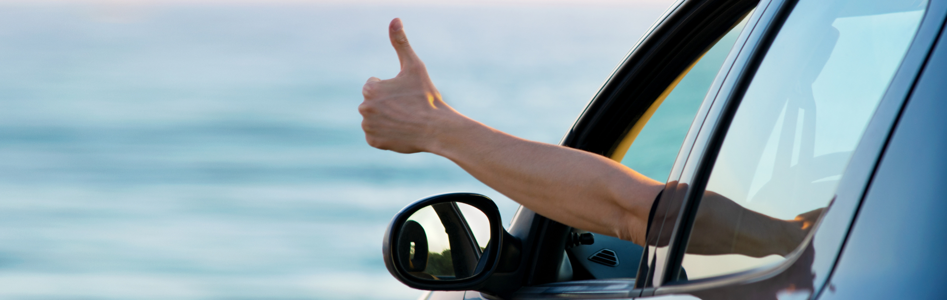 Driver in a car by the ocean displaying a thumbs up out his driver window