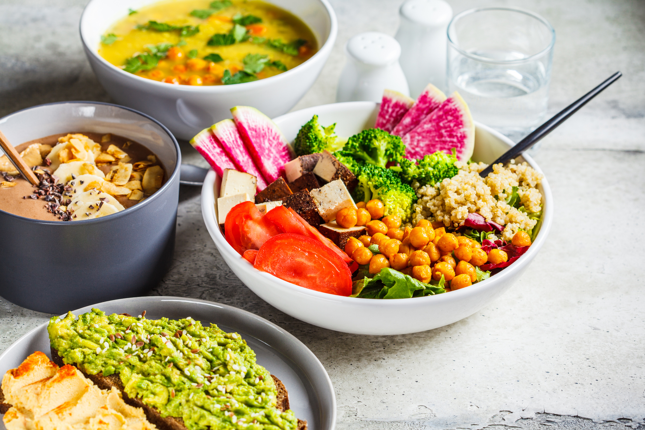 Vegan lunch. Chocolate smoothie bowl, Buddha bowl with tofu, chickpeas and quinoa, lentil soup and toasts on a gray background.