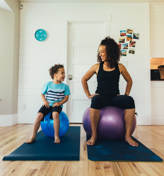 A mother exercises on a fitness ball with her young son inside their home. She is teaching the boy the importance of a healthy lifestyle by proper stretching and exercising