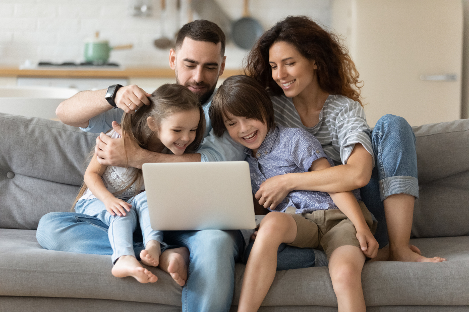 A young smiling family of four sitting on a couch, looking at a laptop