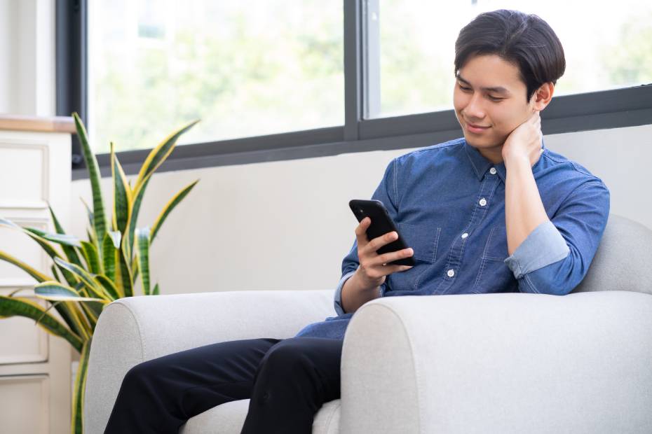 A young man in his 20s in a blue shirt sitting on a white couch, looking at his smartphone