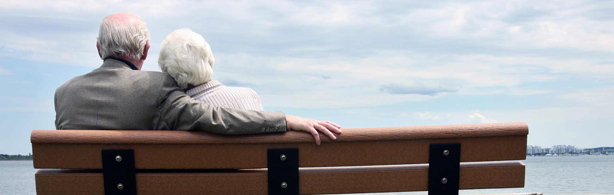 An elderly couple sitting together on a bench, looking out onto the waterfront.